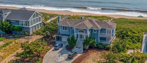 A coastal house with a large driveway, two cars parked, and a view of the ocean.