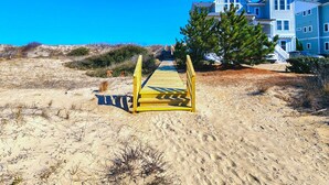 A sandy beach with a wooden boardwalk leading to a row of blue houses. - Luap's Place - Oceanside 5 BR home w/ Private Pool and Hot Tub (Corolla)