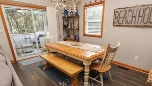 A dining area with a wooden table, bench, and chairs. There is a bookshelf with books and decorative items. A window with blinds and a sign that reads "BEACH HOUSE" are visible. - Luap's Place - Oceanside 5 BR home w/ Private Pool and Hot Tub (Corolla)