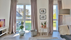A modern living room with large windows, a flat-screen TV, a floor lamp, and a small table with potted plants.