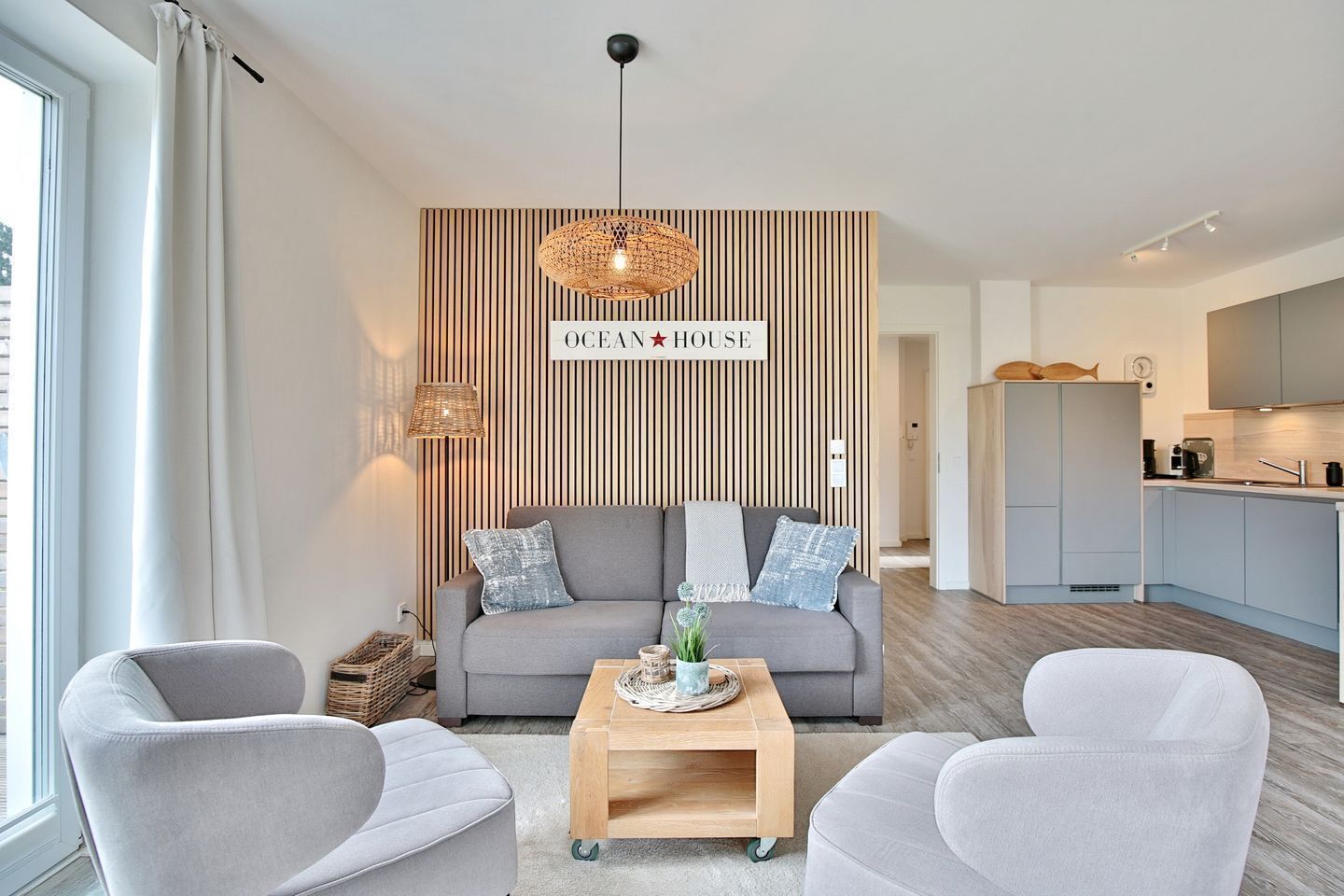 Ein modernes Wohnzimmer mit einem grauen Sofa, zwei Sesseln, einem Holztisch und einem an der Wand befestigten Schild mit der Aufschrift 'OCEAN HOUSE'.