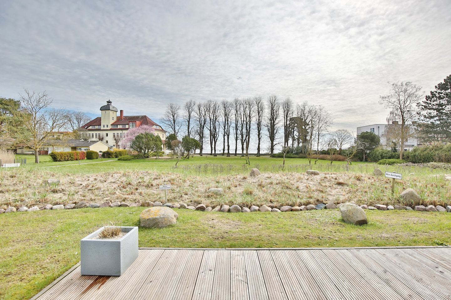 Um jardim ajardinado com um deck de madeira, um caminho de pedra e um edifício com telhado vermelho ao fundo.