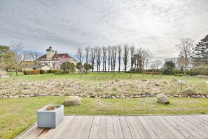 Un jardín ajardinado con una terraza de madera, un sendero de piedra y un edificio con techo rojo al fondo.
