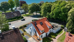 A residential area with houses featuring red-tiled roofs, a central white building with a sloped roof, and a view of a body of water in the background. - Seestern Apartment 3 - Haus am Seehof 7 (Sierksdorf)
