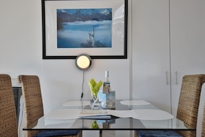 A dining area with a glass table, wicker chairs, a framed picture of a castle, a lit wall lamp, and a bottle of wine.