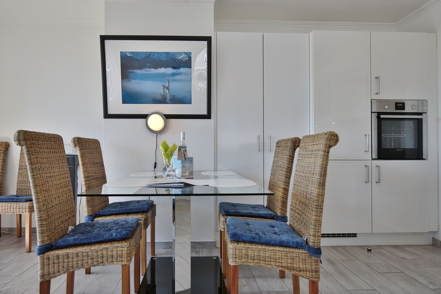 A modern kitchen with white cabinets, a built-in oven, and a dining area with wicker chairs and a glass table.