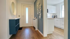Un couloir avec un plancher en bois, une porte blanche, un meuble de rangement bleu et un coin cuisine avec des armoires blanches et une machine à café.