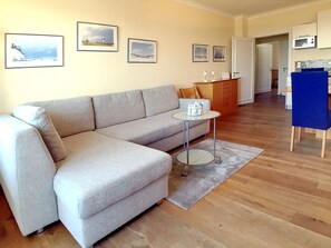 Een woonkamer met een grijze bank, een klein rond tafeltje en een blauwe stoel. Op de muur hangen afbeeldingen in lijsten en er is een keuken in de achtergrond te zien.