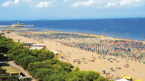 Une plage avec de nombreux parasols et beaucoup de monde, une jetée en arrière-plan et un ciel dégagé.