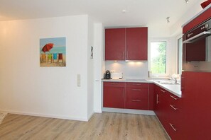 A modern kitchen with red cabinets, a white countertop, and a window providing natural light. - Kurparkresidenz Apartment 25 - Kurparkresidenz (Scharbeutz)