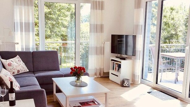 Un moderno salón con un sofá gris, una mesa de centro blanca con un jarrón de flores, un televisor de pantalla plana sobre un mueble blanco, y grandes ventanales que ofrecen vistas a zonas verdes.