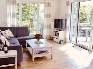 Un salon moderne avec un canapé gris, une table basse blanche ornée d’un vase de fleurs, un téléviseur à écran plat posé sur un meuble blanc, et de grandes fenêtres donnant sur un paysage verdoyant.