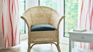 Una silla de mimbre con un cojín oscuro en una habitación con ventanas amplias y un escritorio blanco.