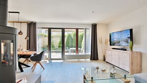 A modern living room with a glass coffee table, a TV mounted on a wooden cabinet, and a sliding glass door leading to an outdoor patio.