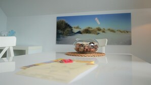 A modern room with a white table, a glass bowl with decorative items, a woven coaster, and a framed beach scene on the wall.