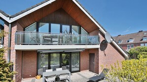 A modern two-story house with a balcony, large windows, and a patio area with outdoor furniture.