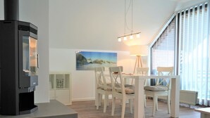 A modern living room with a wood-burning stove, a dining table with chairs, a painting on the wall, and a large window with blinds.