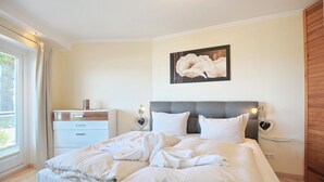 Ett hotellrum med en stor säng, sängbord, ett litet bord, ett fönster med gardiner och ett konstverk som hänger på väggen.
