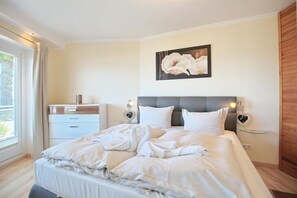 A hotel bedroom with a large bed, bedside tables, a nightstand, a window with curtains, and a wall-mounted artwork.