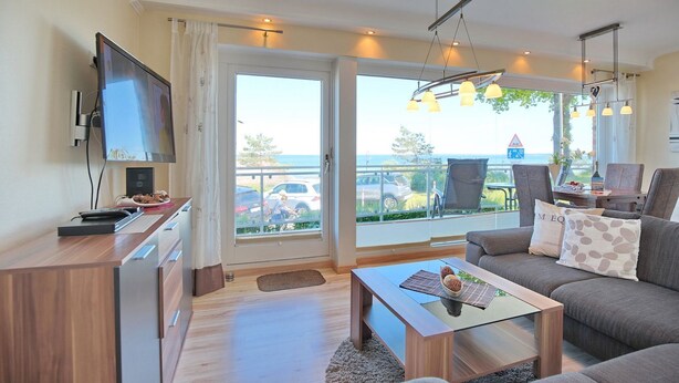 Ett modernt vardagsrum med en soffa, ett soffbord och en tv. Det finns en balkong med utsikt över havet och parkerade bilar.