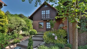 Une maison en bois à deux étages, avec des fondations en briques, un porche à l’avant et un jardin bien entretenu où poussent diverses plantes et fleurs.