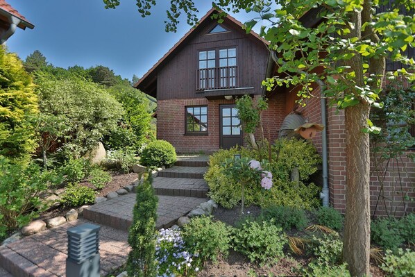 A two-story wooden house with a brick foundation, a front porch, and a well-maintained garden with various plants and flowers.