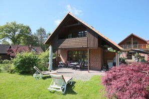 A two-story wooden house with a balcony, a patio with outdoor furniture, and a well-maintained garden.
