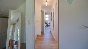 Un couloir doté d’un plancher en bois, de murs blancs et d’une porte donnant sur une pièce où se trouvent une chaise et une fenêtre.