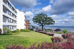 Ein modernes Wohnhaus mit gepflegtem Rasen, einem großen Baum und Blick auf das Meer sowie eine Brücke im Hintergrund.