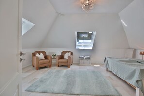 Une chambre avec un plafond en pente, un puits de lumière, deux fauteuils, une table de chevet avec une lampe et un lit recouvert d’une couverture verte.