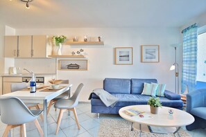 A modern living room with a blue sofa, a dining table with chairs, and a kitchen area with cabinets and a countertop. - Hansa Residence II Apartment 12 - Hansa Residence II (Scharbeutz)