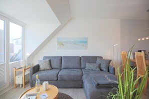 A modern living room with a grey sofa, a small round table, and a painting of a beach scene. - Hansa Residence II Apartment 25 - Hansa Residence II (Scharbeutz)