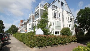A modern multi-story residential building with balconies, a paved sidewalk, and parked cars.