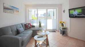 A living room with a grey sofa, a small round table, a TV, and a window with a view of greenery.
