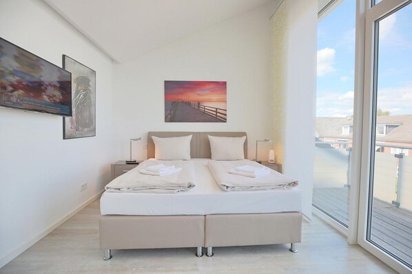 A modern bedroom with a large bed, a nightstand, a bedside table, a window with a view of buildings, and two framed artworks on the wall.