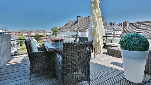 Eine Dachterrasse mit Korbmöbeln, einem Tisch mit Blumen, einer Topfpflanze und Blick auf Gebäude.
