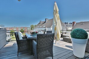 Eine Dachterrasse mit Korbmöbeln, einem Tisch mit Blumen, einer Topfpflanze und Blick auf Gebäude.