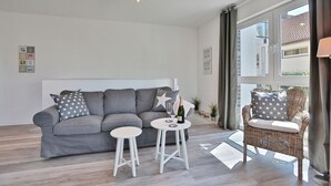 Ein modernes Wohnzimmer mit einem grauen Sofa, zwei weißen Hocker, einem Korbstuhl und einem großen Fenster mit Vorhängen.