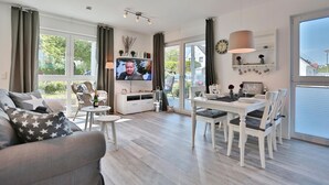 Ein modernes Wohnzimmer mit Flachbildfernseher, einem Essbereich mit weißer Möbel und großen Fenstern mit Blick ins Freie.