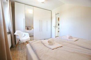 Una habitación de hotel moderna con un armario amplio, una silla blanca, dos camas con toallas y un baño a la vista.
