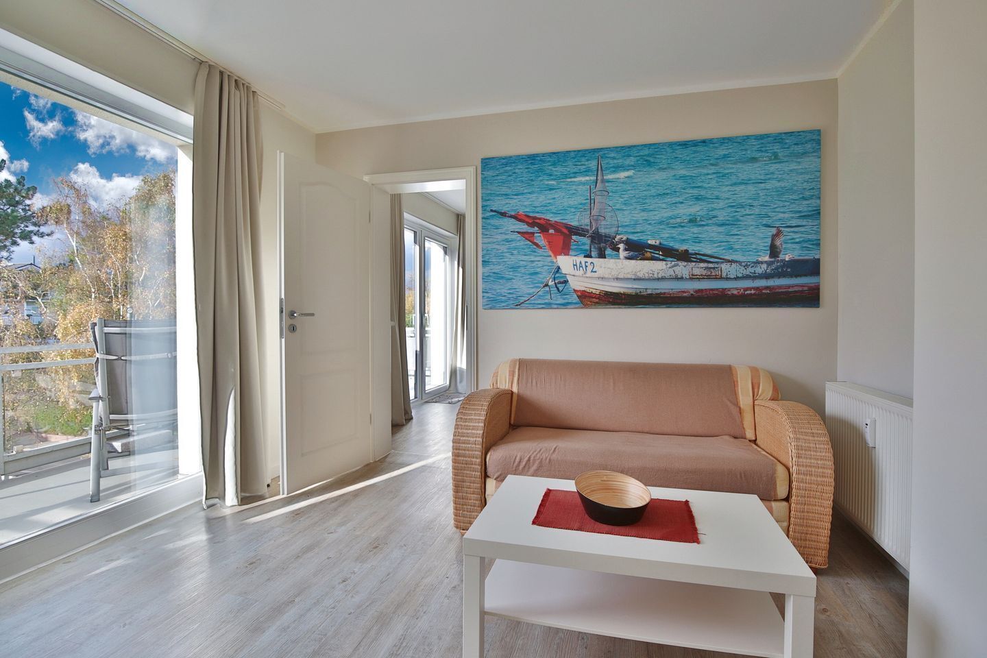 Ein modernes Wohnzimmer mit einer Couch, einem Tisch mit einer Schüssel, einem Gemälde mit einem Boot an der Wand und Blick ins Freie durch ein großes Fenster.