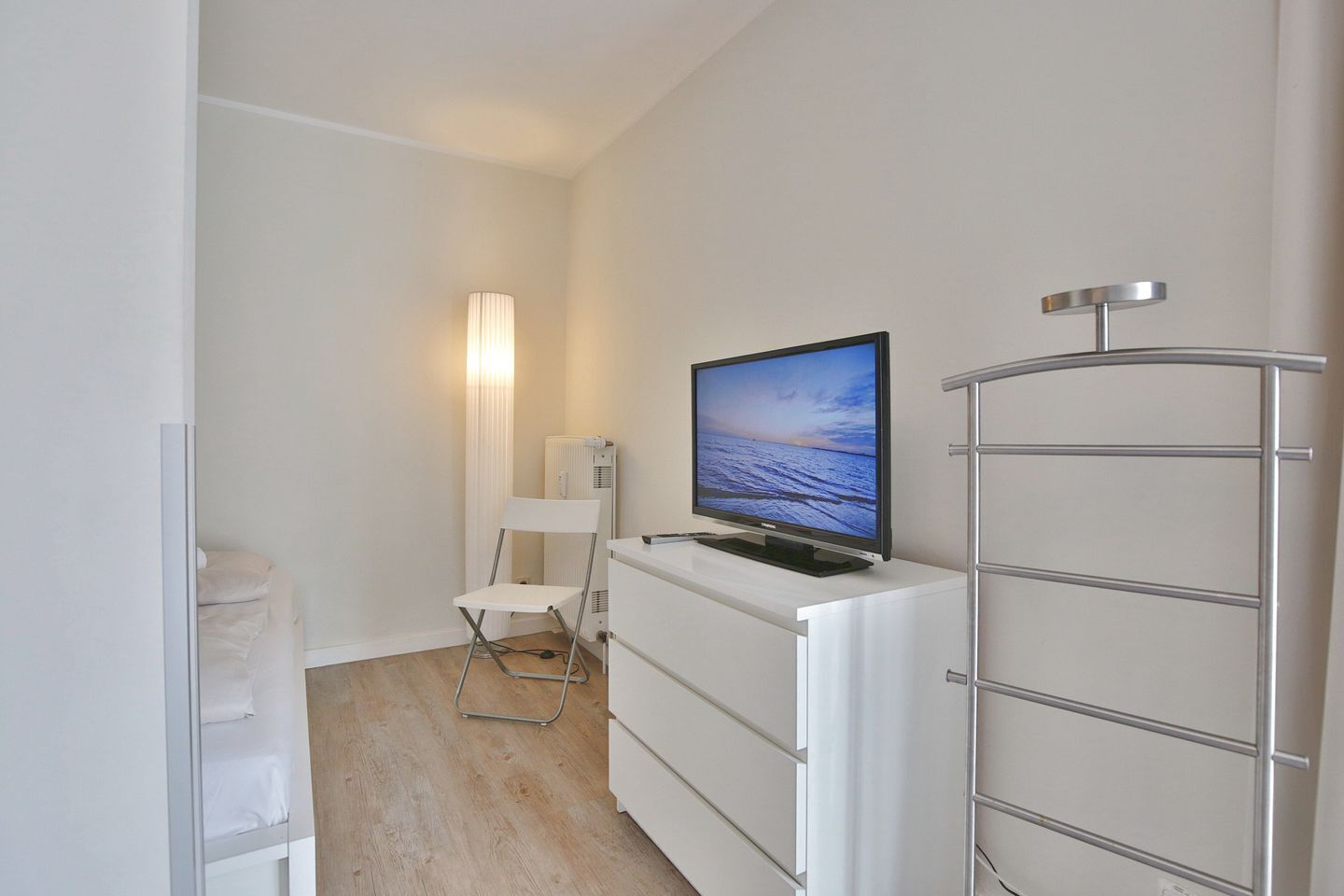 A small, modern room with a flat-screen TV on a white dresser, a white chair, and a metal radiator.