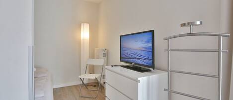 A small, modern room with a flat-screen TV on a white dresser, a white chair, and a metal radiator.