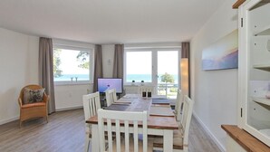 Un comedor con una larga mesa, sillas blancas y un televisor. Hay ventanas con cortinas, vista al mar y un cuadro en la pared.