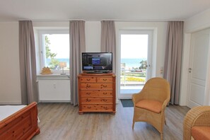 Een kamer met een televisie op een houten dressoir, een rieten stoel en uitzicht op de zee door grote ramen.