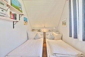 Quarto com duas camas de solteiro, uma estante com livros e uma pintura, uma escrivaninha com abajur e uma janela com cortinas.