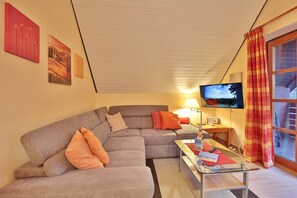 Un salón con un sofá gris, cojines naranjas, una mesa de centro de cristal, un televisor de pantalla plana y una ventana con cortina roja y amarilla.