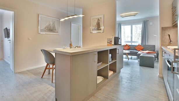 A modern kitchen with a central island, a dining area with a sofa and a coffee table, and a living room with a large window.