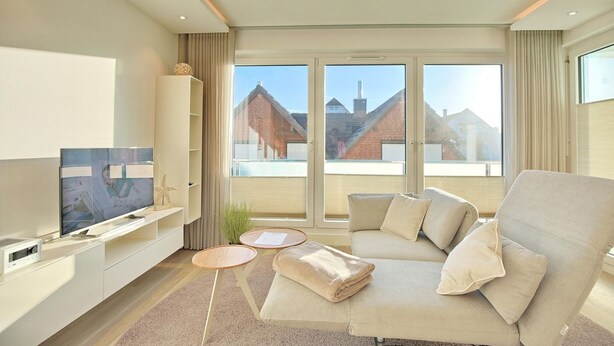 Ett modernt vardagsrum med en stor platt-TV, en beige soffa och utsikt över byggnader genom stora fönster.