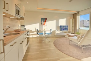 Een moderne keuken met een houten aanrecht, een inbouwoven en een spoelbak. Er is een eethoek met een tafel en stoelen, en een bank in de woonkamer.